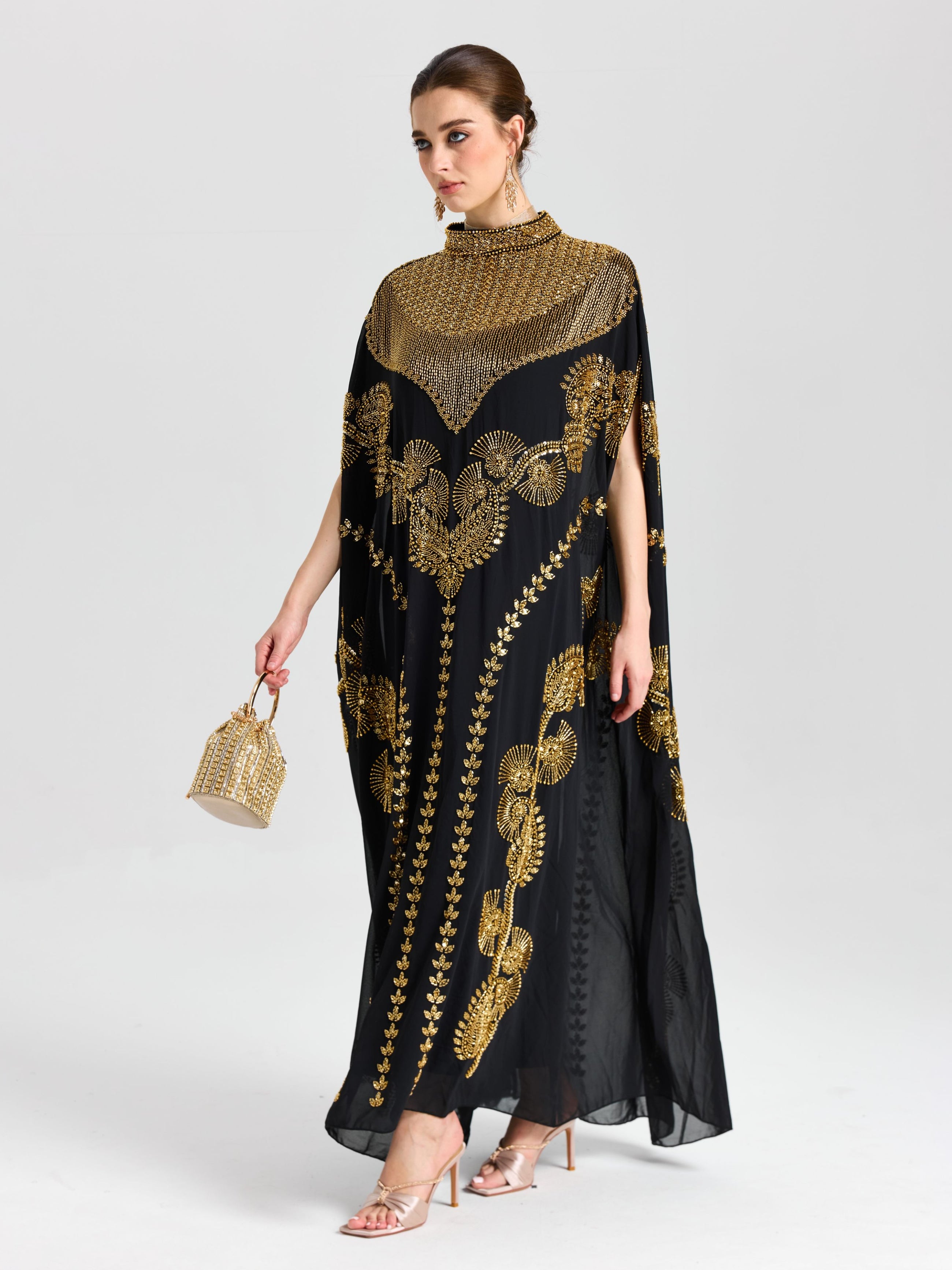 A woman wearing Social Queen’s Beaded Embroidered Cape Dress—featuring Persian-inspired embroidery, a high collar, chiffon cape sleeves—paired with a beaded handbag and heeled sandals for an elegant evening look.