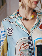 A woman wears the Social Queen Abstract Print Mulberry Silk Shirt, featuring a vibrant resort-inspired pattern, styled with a beaded necklace and oversized dangling earrings; only her lower face and upper torso are shown.