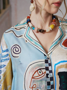 A woman wears the Social Queen Abstract Print Mulberry Silk Shirt, featuring a vibrant resort-inspired pattern, styled with a beaded necklace and oversized dangling earrings; only her lower face and upper torso are shown.