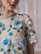 A woman models the Social Queen 3D Floral Cutwork Lace Top, featuring sheer guipure lace with blue and yellow floral embroidery, paired with a pearl necklace and earring; the image is cropped from chin to upper arm against a textured background.