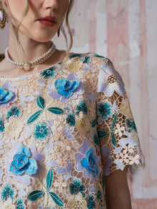A woman models the Social Queen 3D Floral Cutwork Lace Top, featuring sheer guipure lace with blue and yellow floral embroidery, paired with a pearl necklace and earring; the image is cropped from chin to upper arm against a textured background.