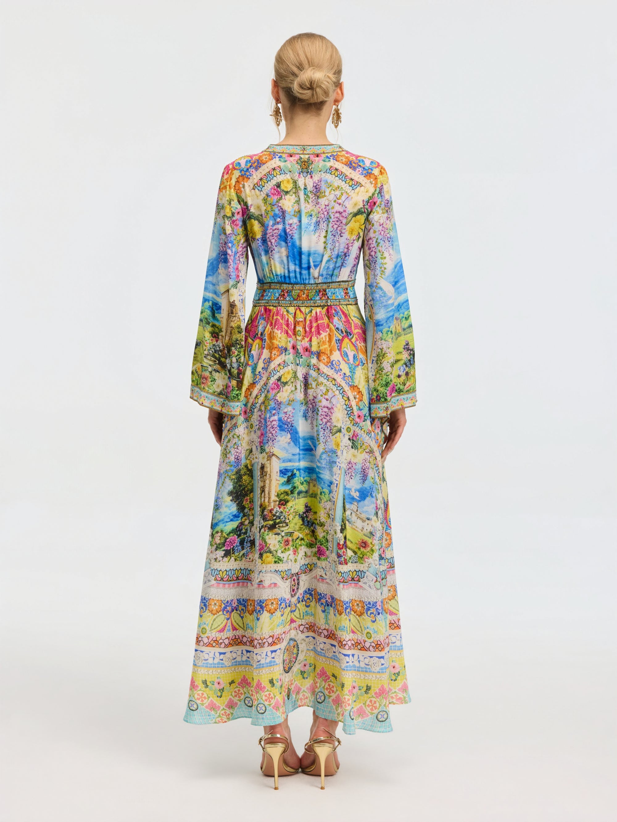 A woman faces away in the Social Queen Garden Print Silk Maxi Dress, featuring long sleeves and vibrant colors, styled with high heels and her hair in a neat bun.