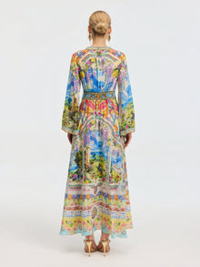 A woman faces away in the Social Queen Garden Print Silk Maxi Dress, featuring long sleeves and vibrant colors, styled with high heels and her hair in a neat bun.