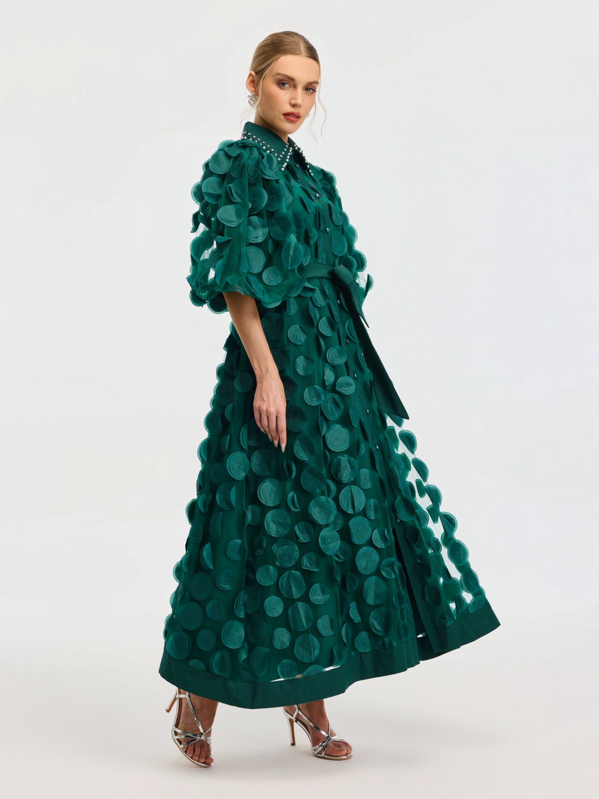 A woman stands against a plain light background in the Social Queen Pearl Embellished Appliqué Dress, an ankle-length green dress with large circular appliqués, a studded collar, and heeled sandals.