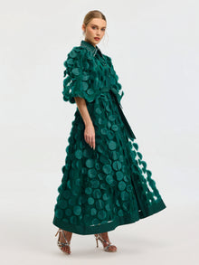 A woman stands against a plain light background in the Social Queen Pearl Embellished Appliqué Dress, an ankle-length green dress with large circular appliqués, a studded collar, and heeled sandals.
