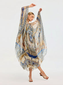 A woman models the Social Queen Byzantine Print Silk Caftan Dress, featuring wide sleeves and a crystal-embellished neckline, standing on neutral heels with arms raised and one leg bent against a plain background.
