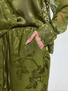 A person wears Social Queen's Botanical Burnout Satin Wide-Leg Pants with a sheer green floral outfit, one hand in the pocket, highlighting detailed sleeve embroidery.