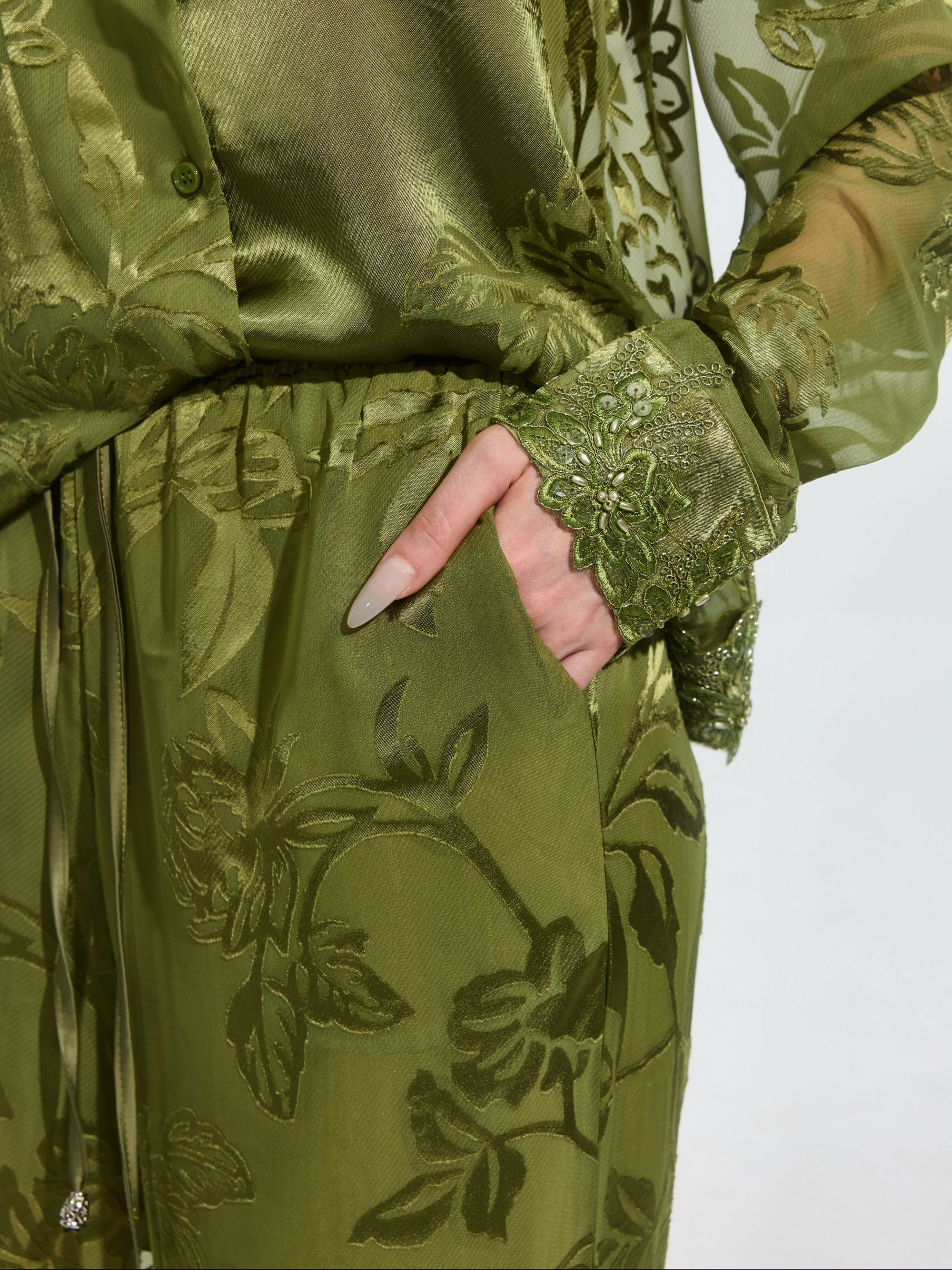 A person wears Social Queen's Botanical Burnout Satin Wide-Leg Pants with a sheer green floral outfit, one hand in the pocket, highlighting detailed sleeve embroidery.