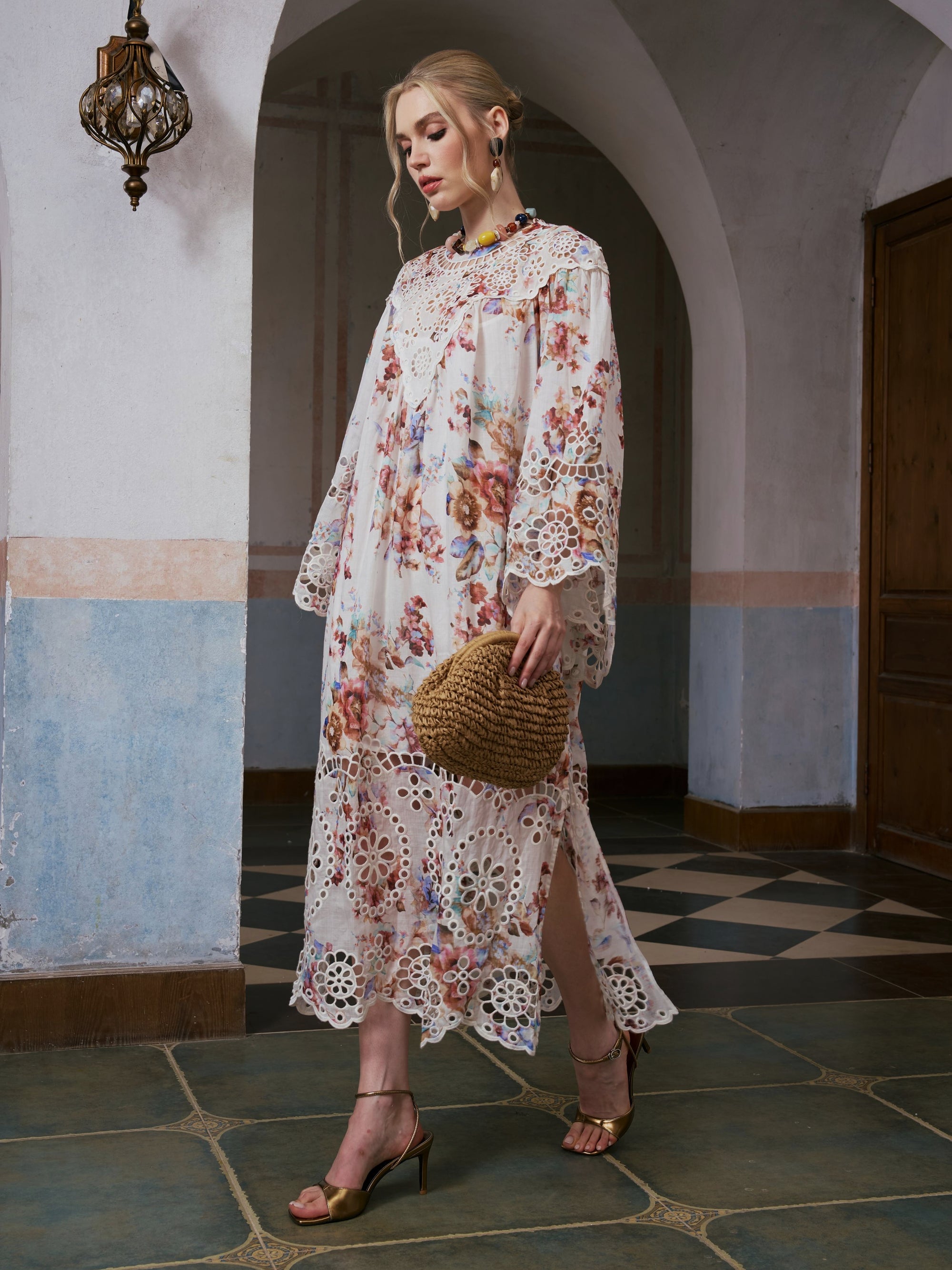 A woman stands in an arched hallway with tiled floors, wearing the Social Queen Floral Print Cutwork Linen Dress—a long maxi dress showcasing a watercolor floral pattern and delicate cutwork embroidery. She carries a round straw bag.