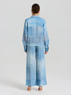 A person stands facing away, wearing the Social Queen Washed Silk Lace Detail Shirt in light blue with lace trim and matching wide-leg pants, paired with beige high-heeled sandals.