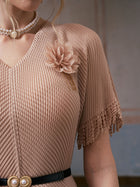 A person wearing the Social Queen Asymmetric Fringe Midi Dress (belt included) in beige, featuring pleated fabric, asymmetrical fringe sleeves, a 3D floral appliqué brooch, and styled with a pearl choker necklace.