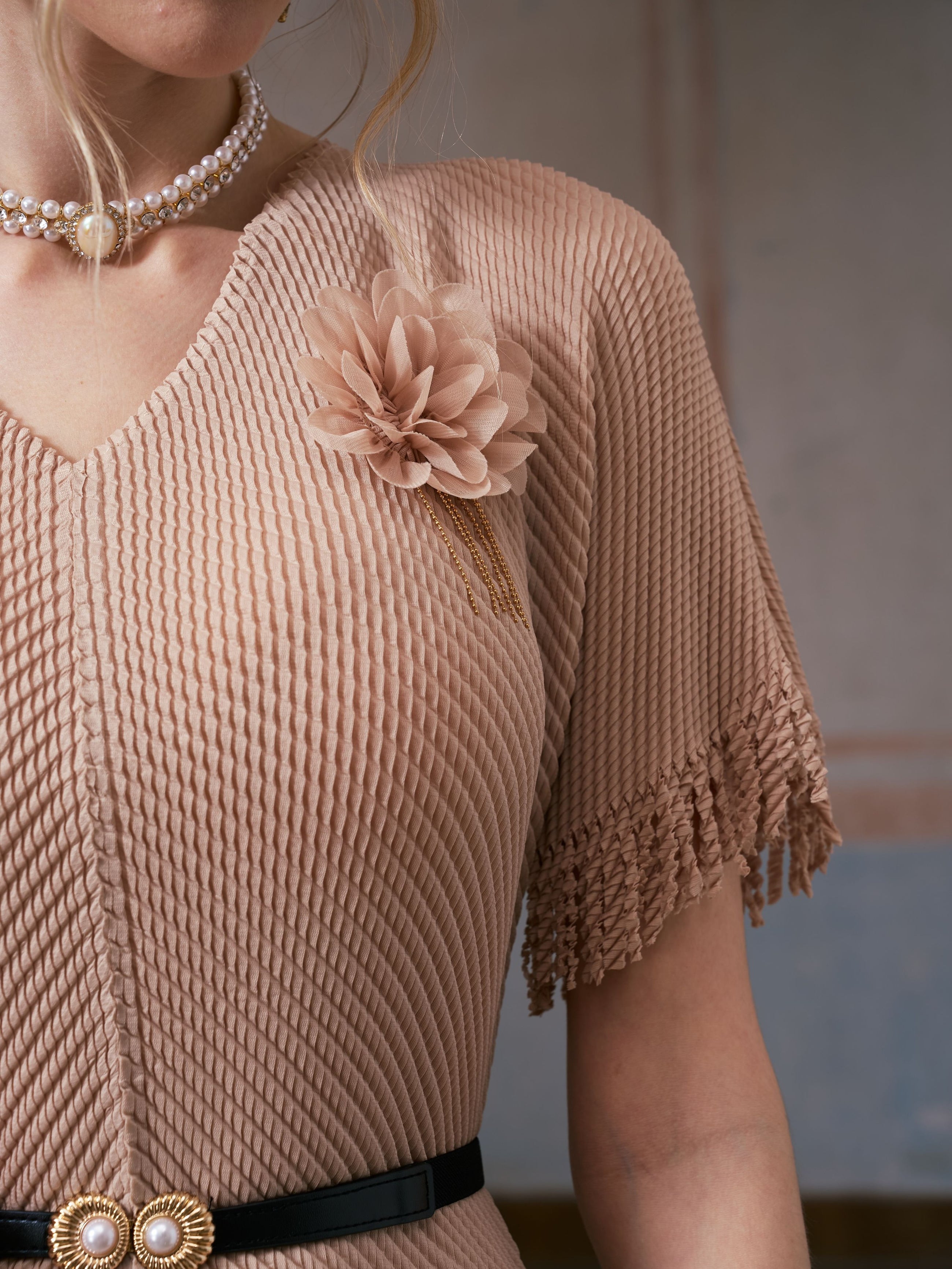 A person wearing the Social Queen Asymmetric Fringe Midi Dress (belt included) in beige, featuring pleated fabric, asymmetrical fringe sleeves, a 3D floral appliqué brooch, and styled with a pearl choker necklace.