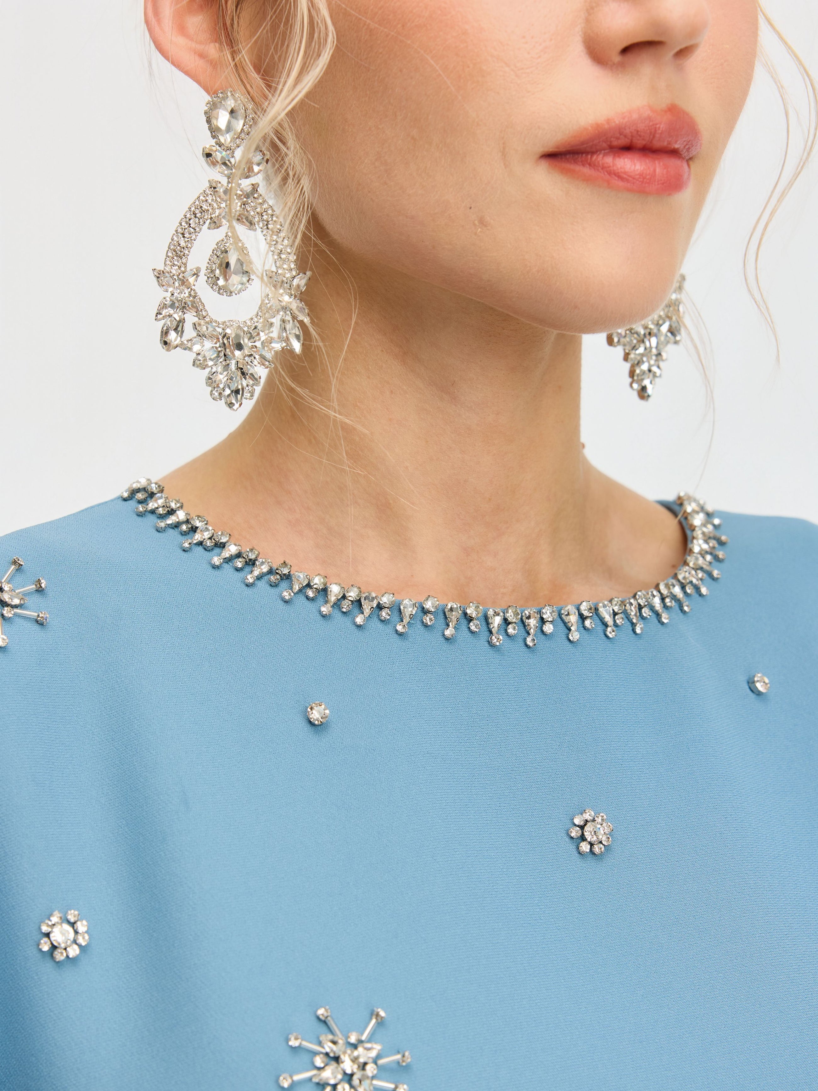 A woman wears the Social Queen Beaded Crystal Embellished Dress, featuring light blue fabric with sparkling crystal details, paired with large silver earrings.