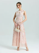 A woman wears the Social Queen Gold Floral Embroidered Dress—a sleeveless, floor-length light pink A-line gown—with sandals, jewelry, subtle makeup, and a small beige purse, standing against a plain light background.