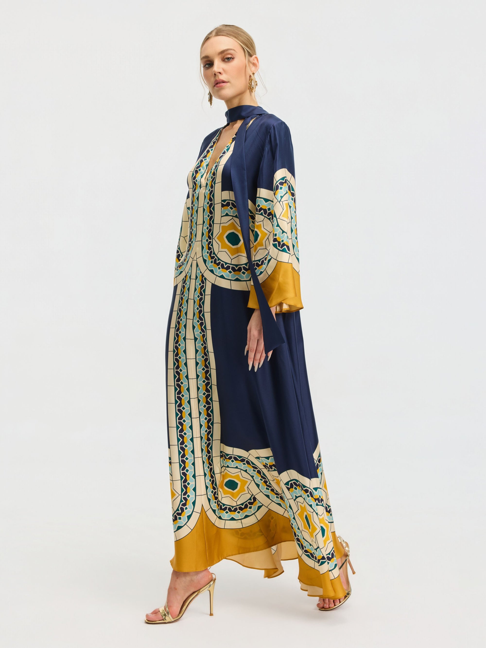 A woman stands sideways in high-heeled sandals, wearing the Social Queen Moroccan Print Silk Caftan Dress—navy and gold with geometric patterns—against a plain light background.