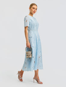 A woman wearing the Social Queen Floral Cutwork Embroidered Dress—a light blue, short-sleeved lace midi dress with delicate floral embroidery—paired with silver high-heeled sandals and a small beaded handbag, stands against a plain white background.