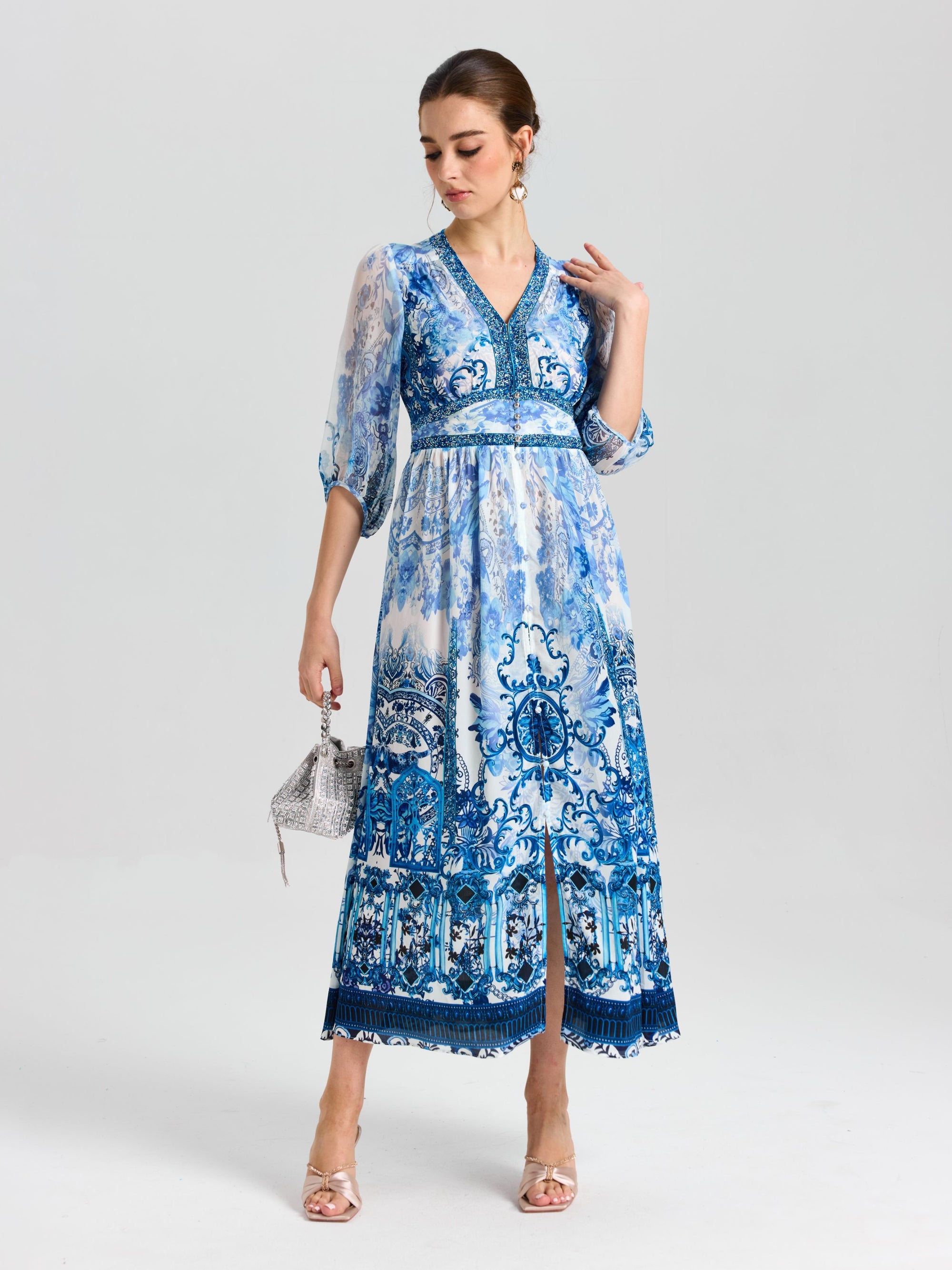 A woman wears the Baroque Placement Print Silk Dress by Social Queen, featuring blue-and-white porcelain-inspired patterns and three-quarter sleeves, holding a silver handbag against a white background.