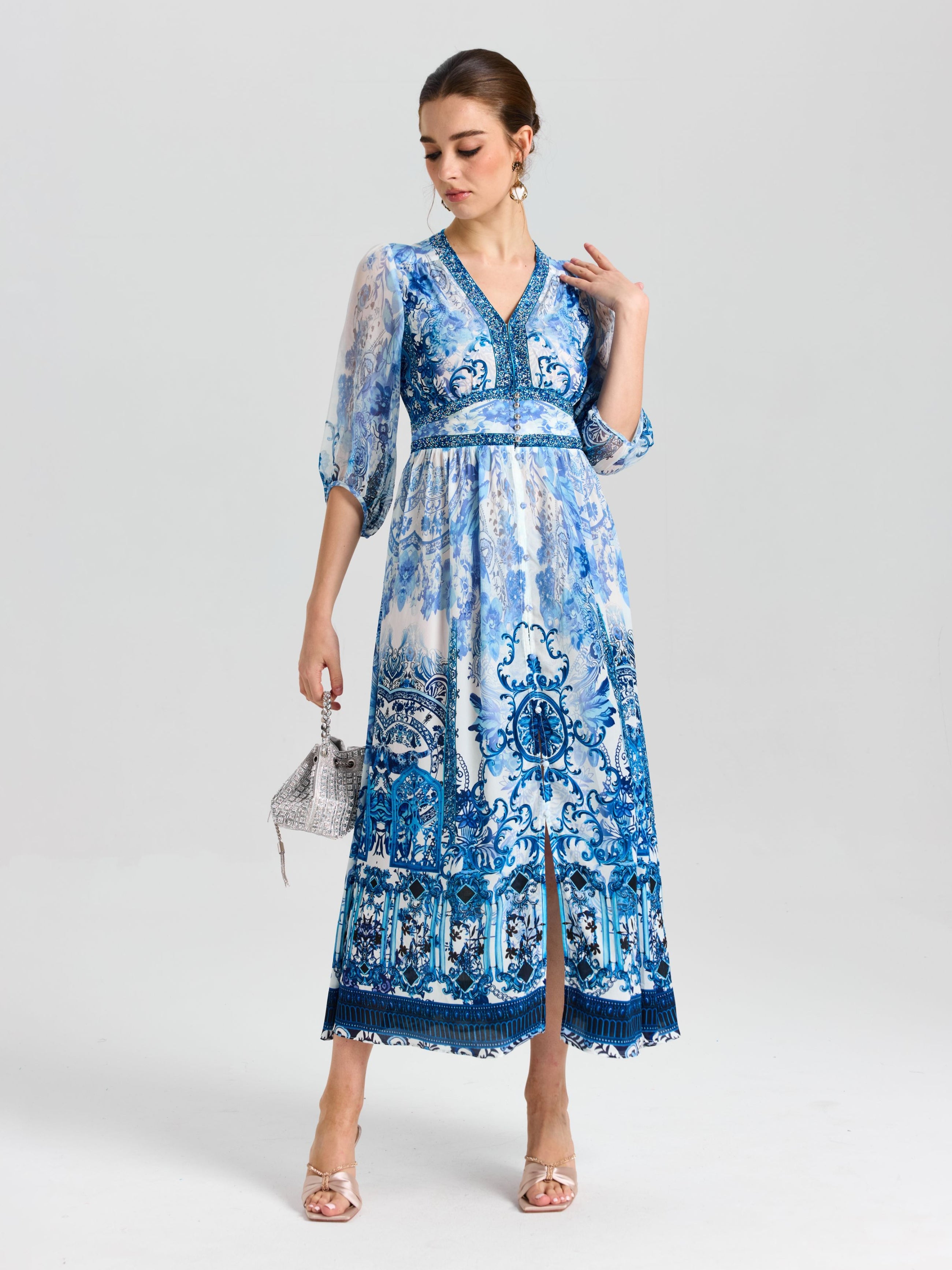 A woman wears the Baroque Placement Print Silk Dress by Social Queen, featuring blue-and-white porcelain-inspired patterns and three-quarter sleeves, holding a silver handbag against a white background.