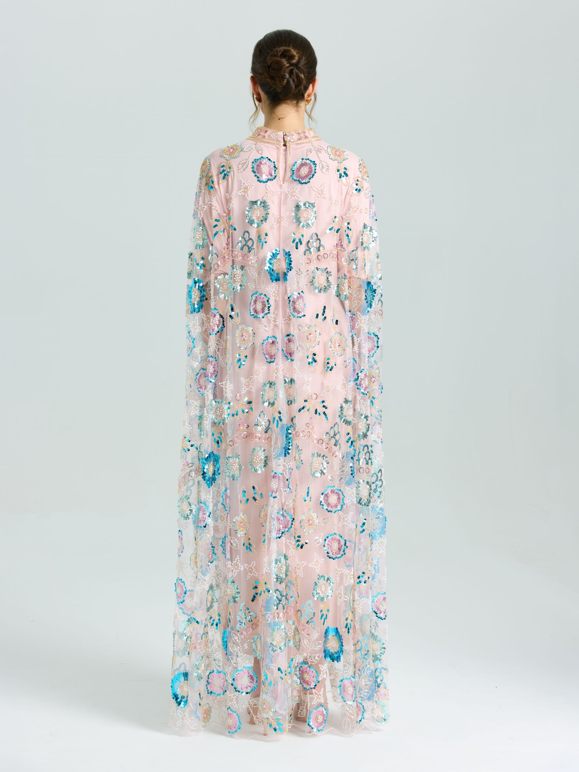 A person faces away in the Social Queen Beaded Floral Embellished Cape Gown, featuring sheer pink fabric with multicolored floral beading and sequins, set against a plain light background.