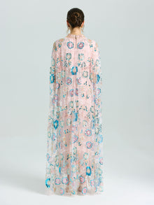 A person faces away in the Social Queen Beaded Floral Embellished Cape Gown, featuring sheer pink fabric with multicolored floral beading and sequins, set against a plain light background.