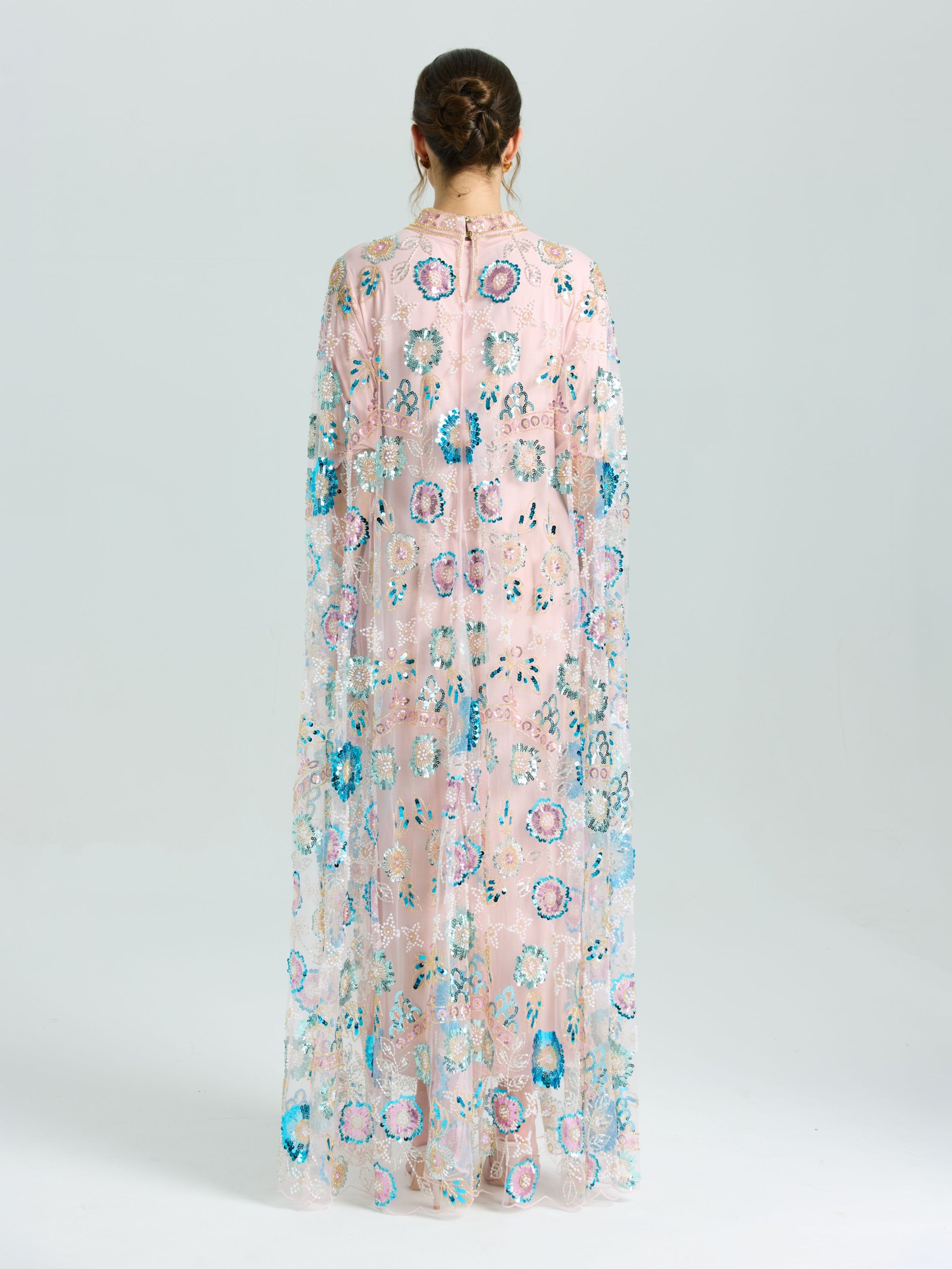 A person faces away in the Social Queen Beaded Floral Embellished Cape Gown, featuring sheer pink fabric with multicolored floral beading and sequins, set against a plain light background.