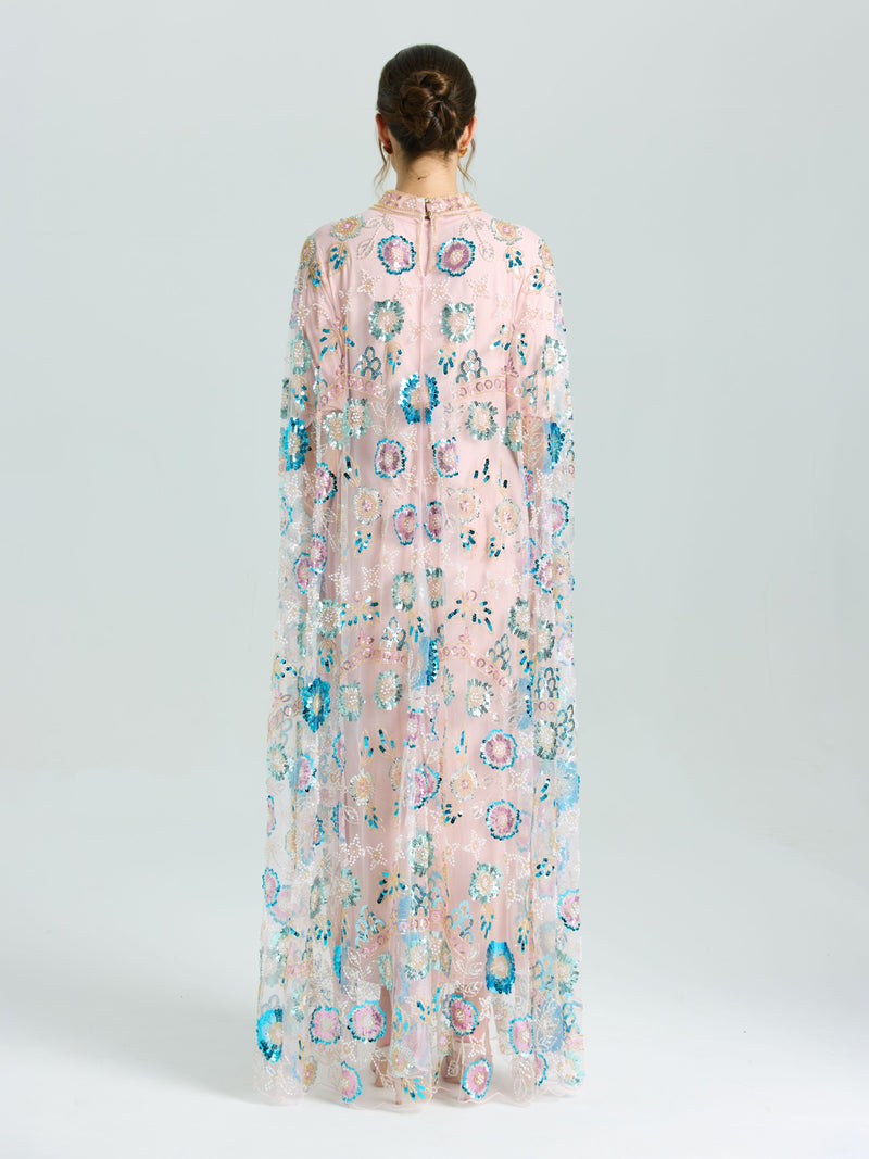 A person faces away in the Social Queen Beaded Floral Embellished Cape Gown, featuring sheer pink fabric with multicolored floral beading and sequins, set against a plain light background.