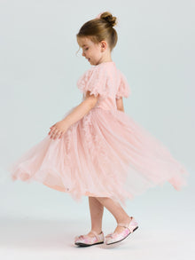 A young girl twirls sideways in a Social Queen Girls Sequin Embellished Tulle Dress, her hair in a bun and floral-patterned shoes completing the look against a plain, light background.