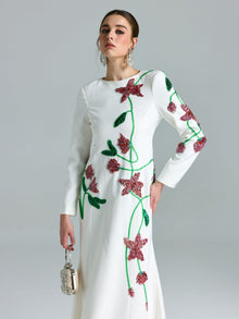 A woman wears the Social Queen Floral Beaded A-Line Gown—a white, long-sleeve dress with red and green floral embellishments—while holding a metallic purse against a light gray background.