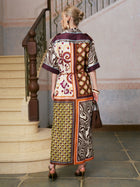 A woman with blonde hair in a bun is seen from behind, standing on a tiled floor near a staircase, wearing the Social Queen Patchwork Print Cotton Shirt Dress (belt included) paired with heels and carrying a brown handbag.