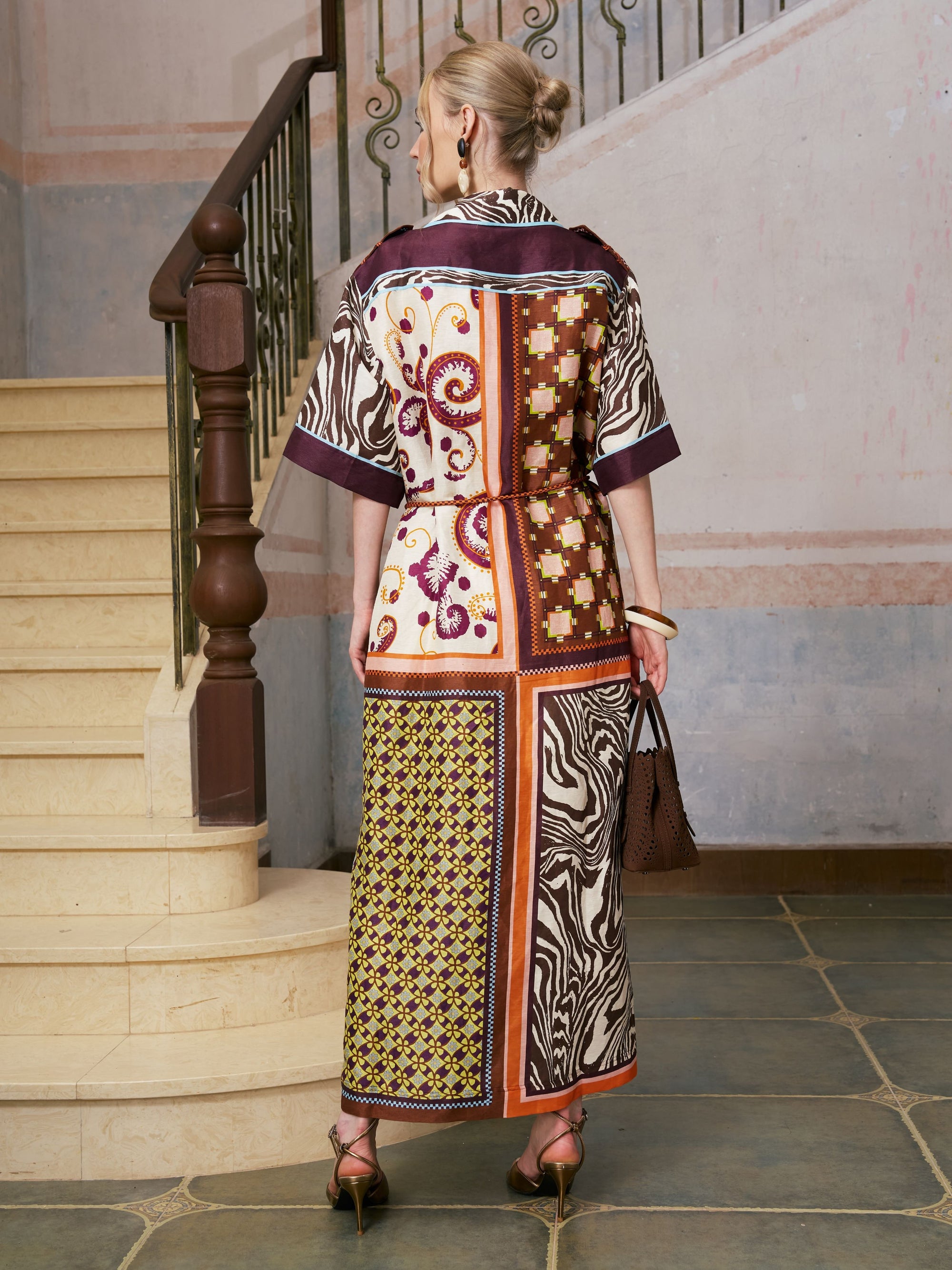 A woman with blonde hair in a bun is seen from behind, standing on a tiled floor near a staircase, wearing the Social Queen Patchwork Print Cotton Shirt Dress (belt included) paired with heels and carrying a brown handbag.