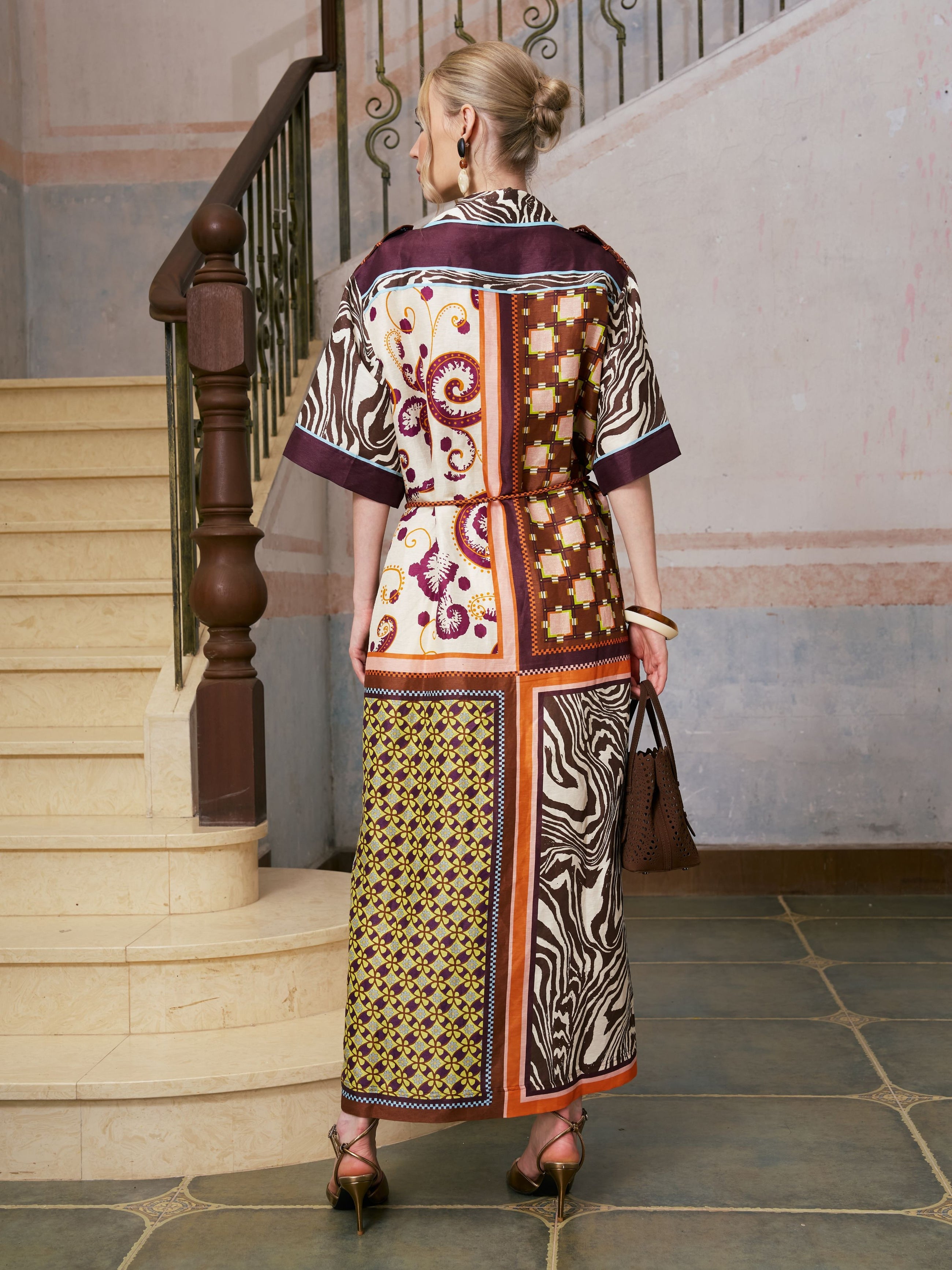 A woman with blonde hair in a bun is seen from behind, standing on a tiled floor near a staircase, wearing the Social Queen Patchwork Print Cotton Shirt Dress (belt included) paired with heels and carrying a brown handbag.