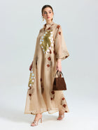 A woman in the Social Queen Placement Embroidered Linen Dress, featuring gold and white floral details and subtle sequins, holds a brown handbag and wears heeled sandals, standing against a plain background.