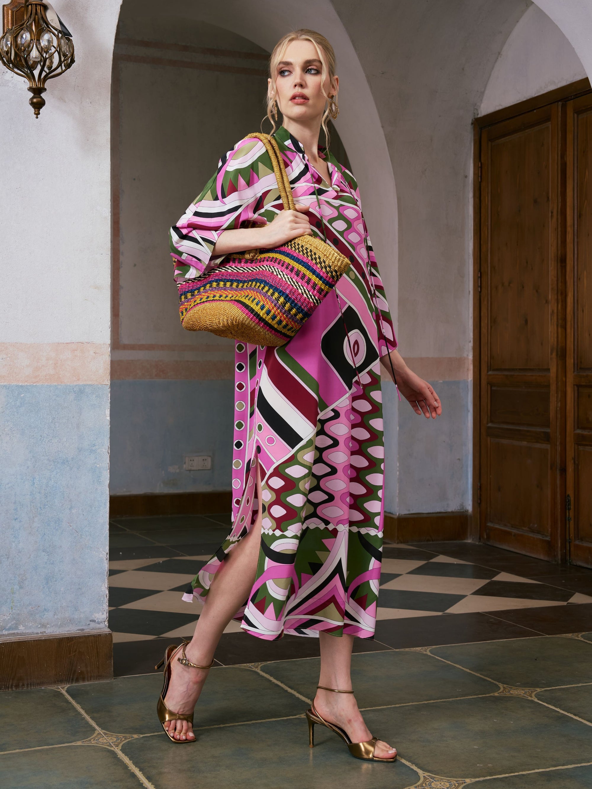 A woman stands indoors wearing the Social Queen Geometric Print Mulberry Silk Dress with a robe-style silhouette, paired with gold heels and carrying a large woven handbag.
