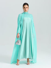 A woman in the Social Queen Beaded Cape Scarf Gown with an A-line silhouette stands against a white background, accessorized with a small silver handbag and matching silver heels.