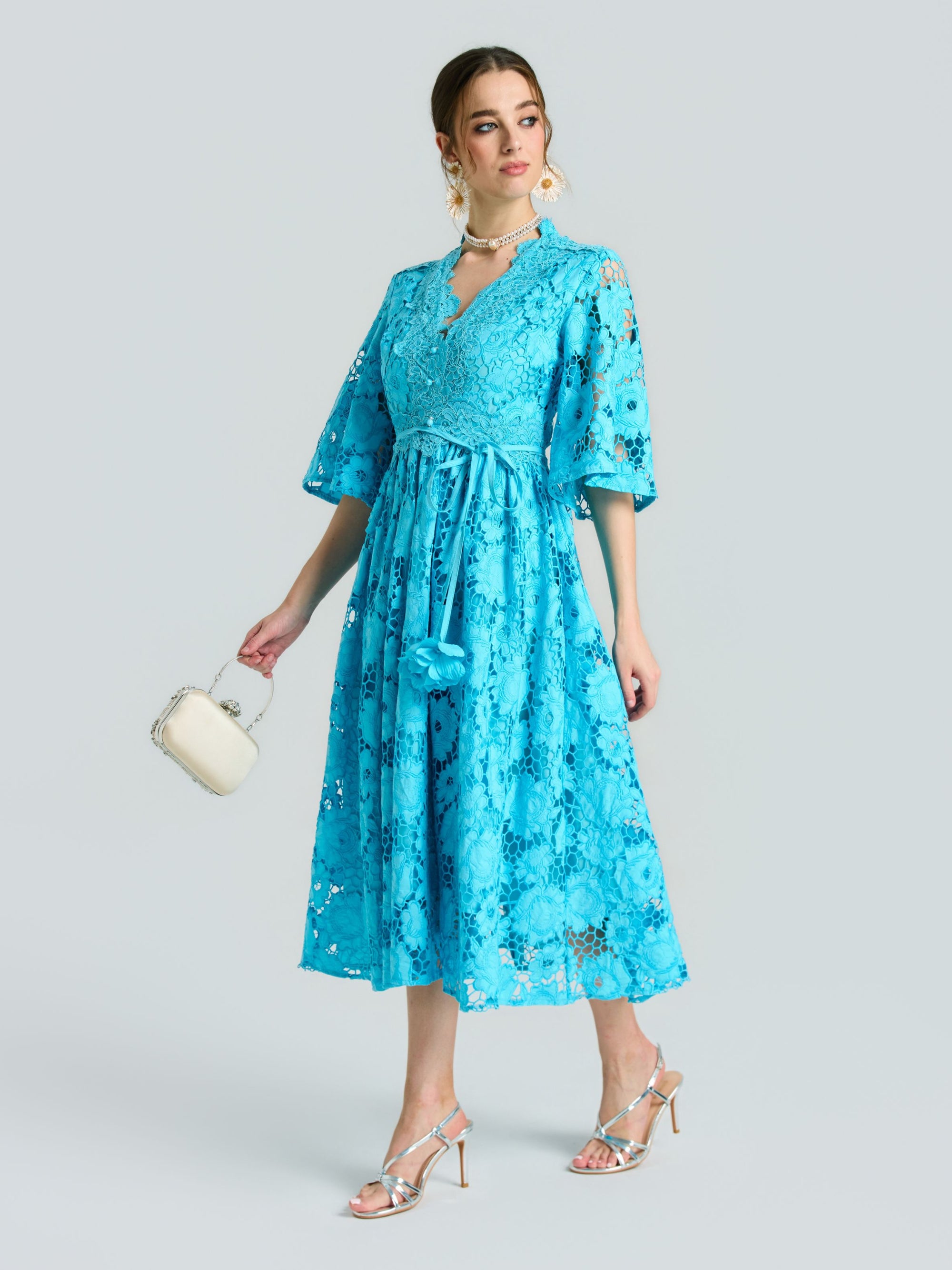 A woman in the Social Queen Rose Cutwork Lace Dress, featuring a bright blue rose-pattern lace, A-line silhouette, and half sleeves, paired with silver high-heeled sandals and a small white bag against a light gray backdrop.