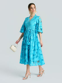 A woman in the Social Queen Rose Cutwork Lace Dress, featuring a bright blue rose-pattern lace, A-line silhouette, and half sleeves, paired with silver high-heeled sandals and a small white bag against a light gray backdrop.