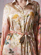 A person wearing the Garden Print Mulberry Silk Shirt Dress by Social Queen, featuring a luxurious, silky finish and a belted waist. Reminiscent of upscale resort wear, this dress showcases bold baroque garden-inspired floral patterns in soft pastel shades.