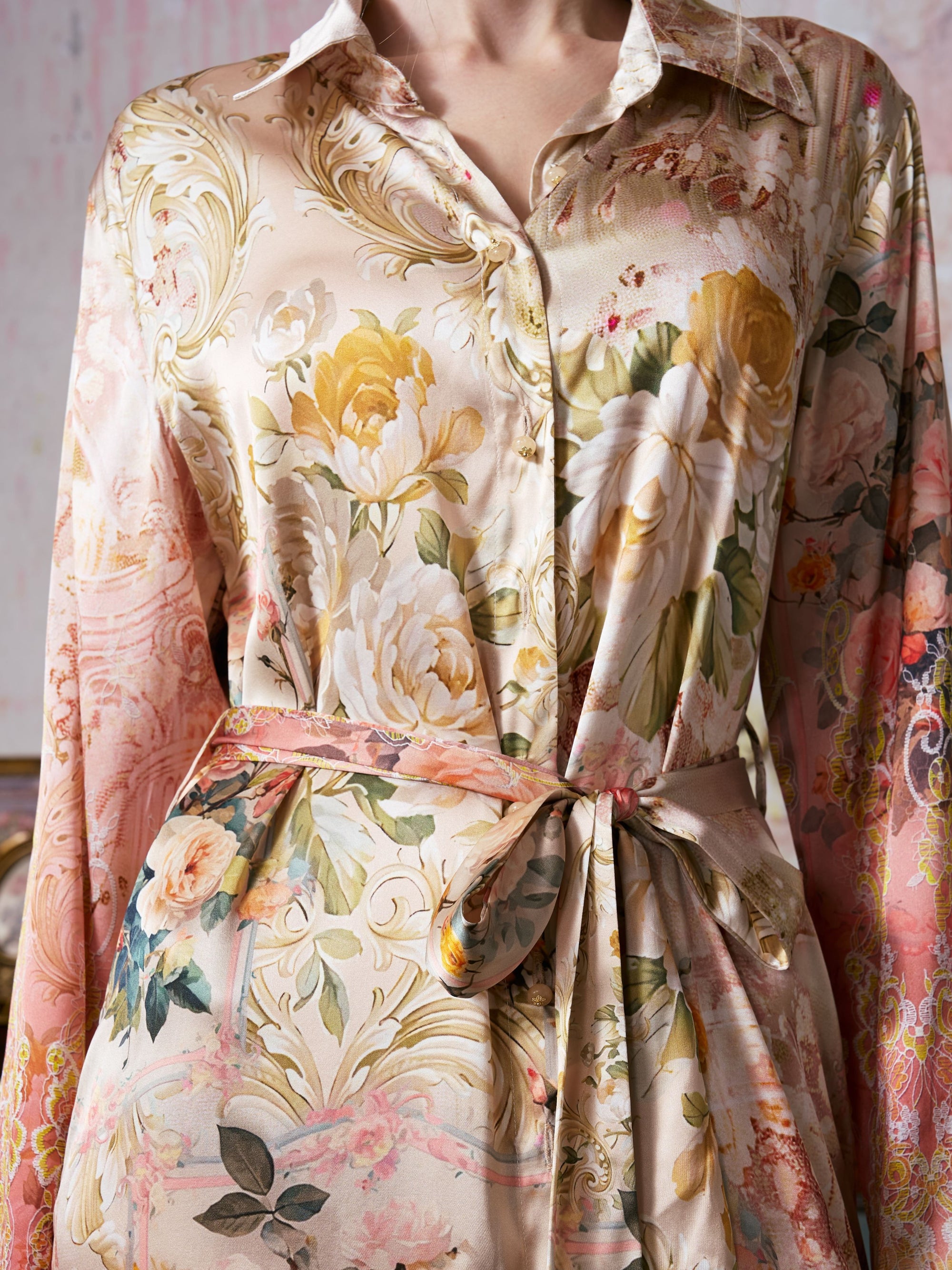 A person wearing the Garden Print Mulberry Silk Shirt Dress by Social Queen, featuring a luxurious, silky finish and a belted waist. Reminiscent of upscale resort wear, this dress showcases bold baroque garden-inspired floral patterns in soft pastel shades.
