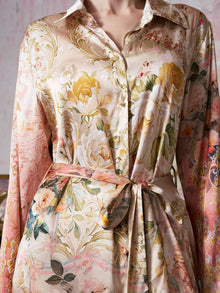 A person wearing the Garden Print Mulberry Silk Shirt Dress by Social Queen, featuring a luxurious, silky finish and a belted waist. Reminiscent of upscale resort wear, this dress showcases bold baroque garden-inspired floral patterns in soft pastel shades.