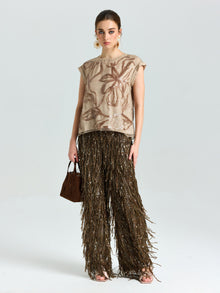 A woman poses against a plain background wearing Social Queen’s Floral Sequin Embroidered Silk Top, styled with brown fringed trousers, a small brown handbag, and chic earrings.