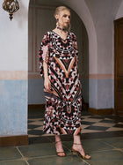 A woman stands indoors wearing the Social Queen Geometric Motif Print Silk Dress (Scarf Included), featuring an elegant mix of pink, black, and beige tones—an elevated resort wear look styled with gold heels and statement earrings.