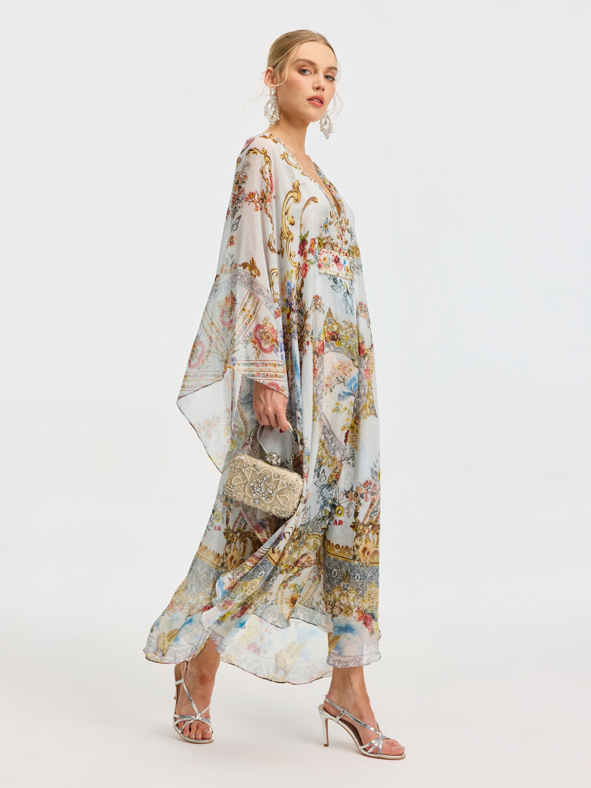 Woman wearing the Social Queen Baroque Floral Print Silk Caftan Dress with sheer sleeves, paired with silver high-heeled sandals and a small ornate handbag against a plain white background.