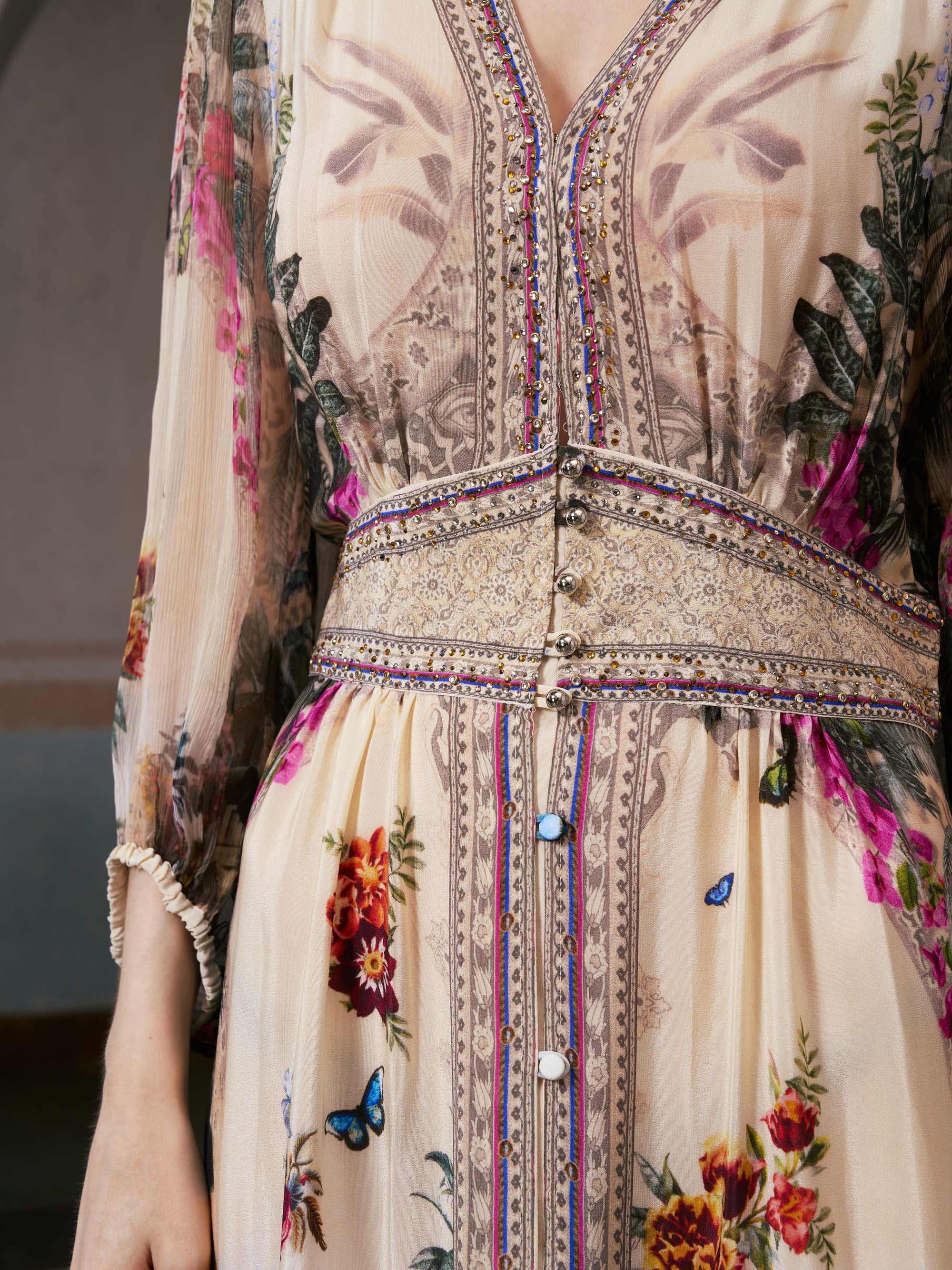 A close-up of a person wearing the Coastal Print Mulberry Silk Dress by Social Queen—a floral, embroidered, long-sleeve dress featuring intricate patterns, buttons, and decorative details at the waist and bodice—offers a refined take on resort wear that evokes effortless elegance.