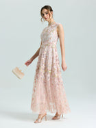 A woman wears the Social Queen Gold Floral Embroidered Dress, a sleeveless, floor-length A-line gown with gold floral details, paired with a beige clutch and matching open-toe heels against a plain background.