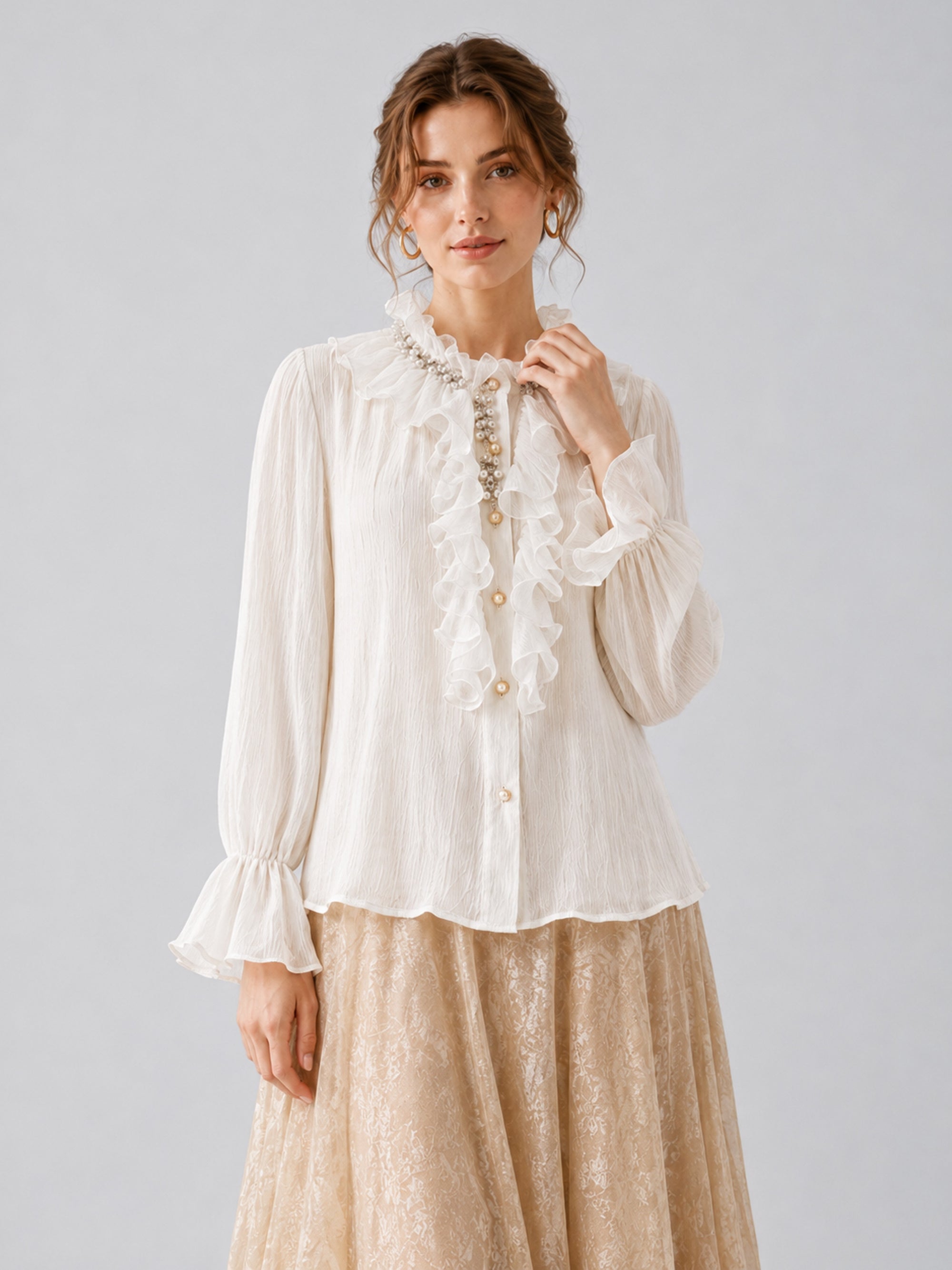 A woman models the Social Queen Pearl Embellished Ruffle Collar Blouse, featuring long sleeves and pearl buttons, paired with a beige lace skirt and large earrings, set against a simple light gray background.