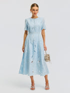 A woman wearing the Social Queen Floral Cutwork Embroidered Dress—a light blue, lace midi dress with short sleeves—holds a small purse while standing against a plain white background.