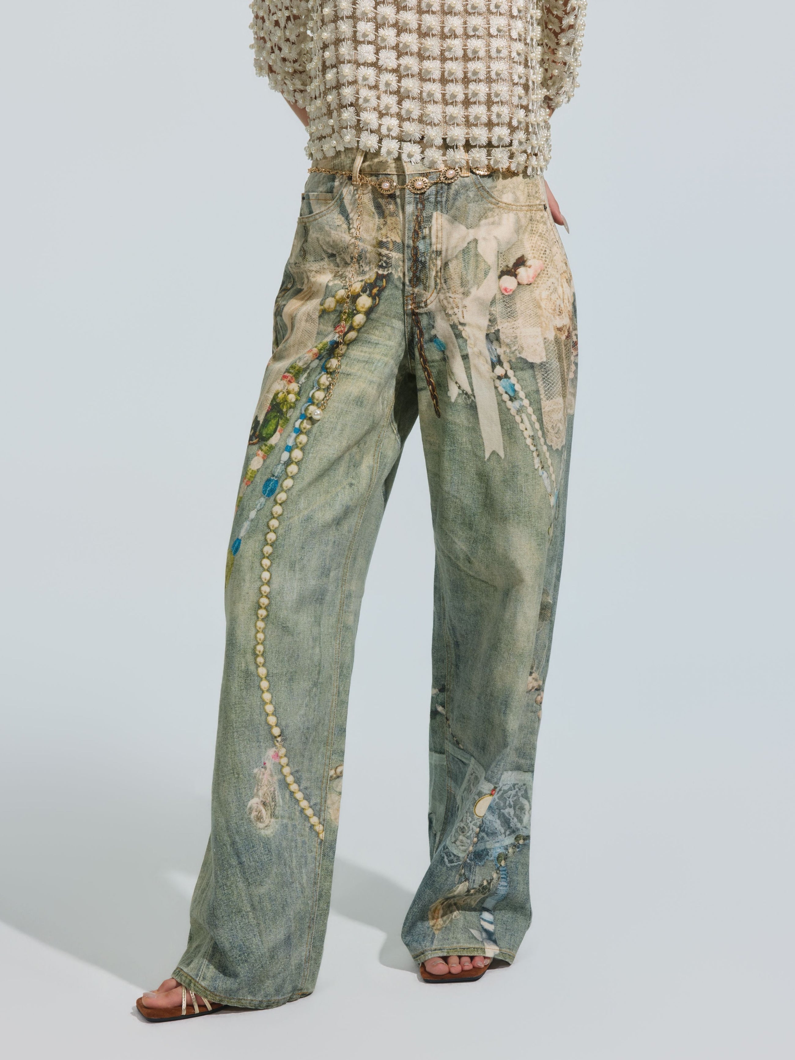A person wears the Social Queen Pearl Chain Printed Jeans—wide-leg, light-wash denim featuring a pearl chain print and bead embellishments—paired with a textured, cream-colored top and open-toe heels. The wide-leg silhouette of the Pearl Chain Printed Jeans creates an effortlessly stylish look, with the upper body partially out of frame.