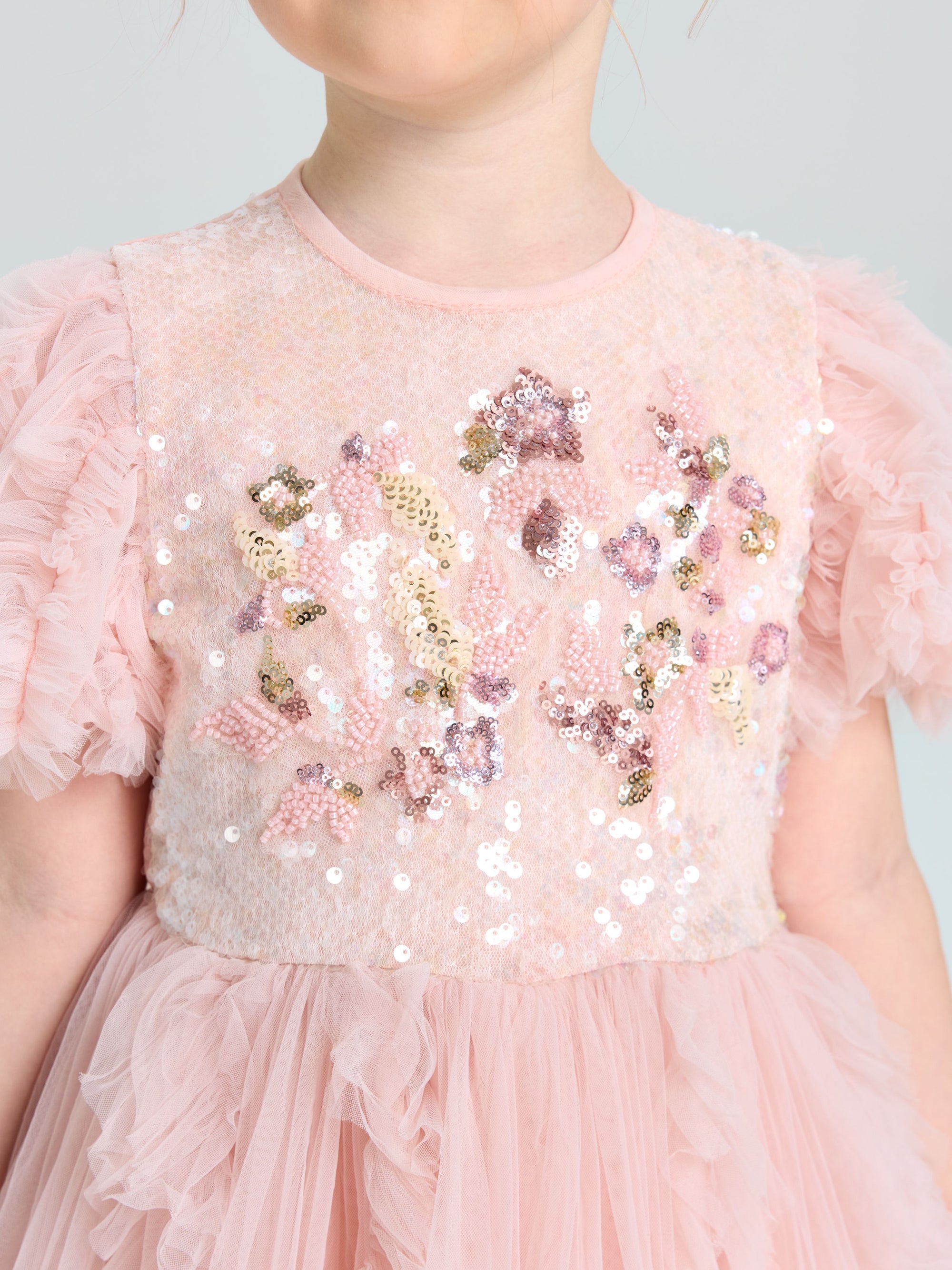A child wears the Social Queen Girls Sequin Embellished Tulle Dress, featuring light pink tulle, ruffled sleeves, sparkling sequins, and floral bodice details.