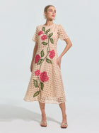 A woman models the Social Queen Hydrangea Embroidered Openwork Dress, a knee-length, short-sleeve beige A-line dress with geometric cutouts and large pink hydrangea and green leaf patterns, set against a plain background.