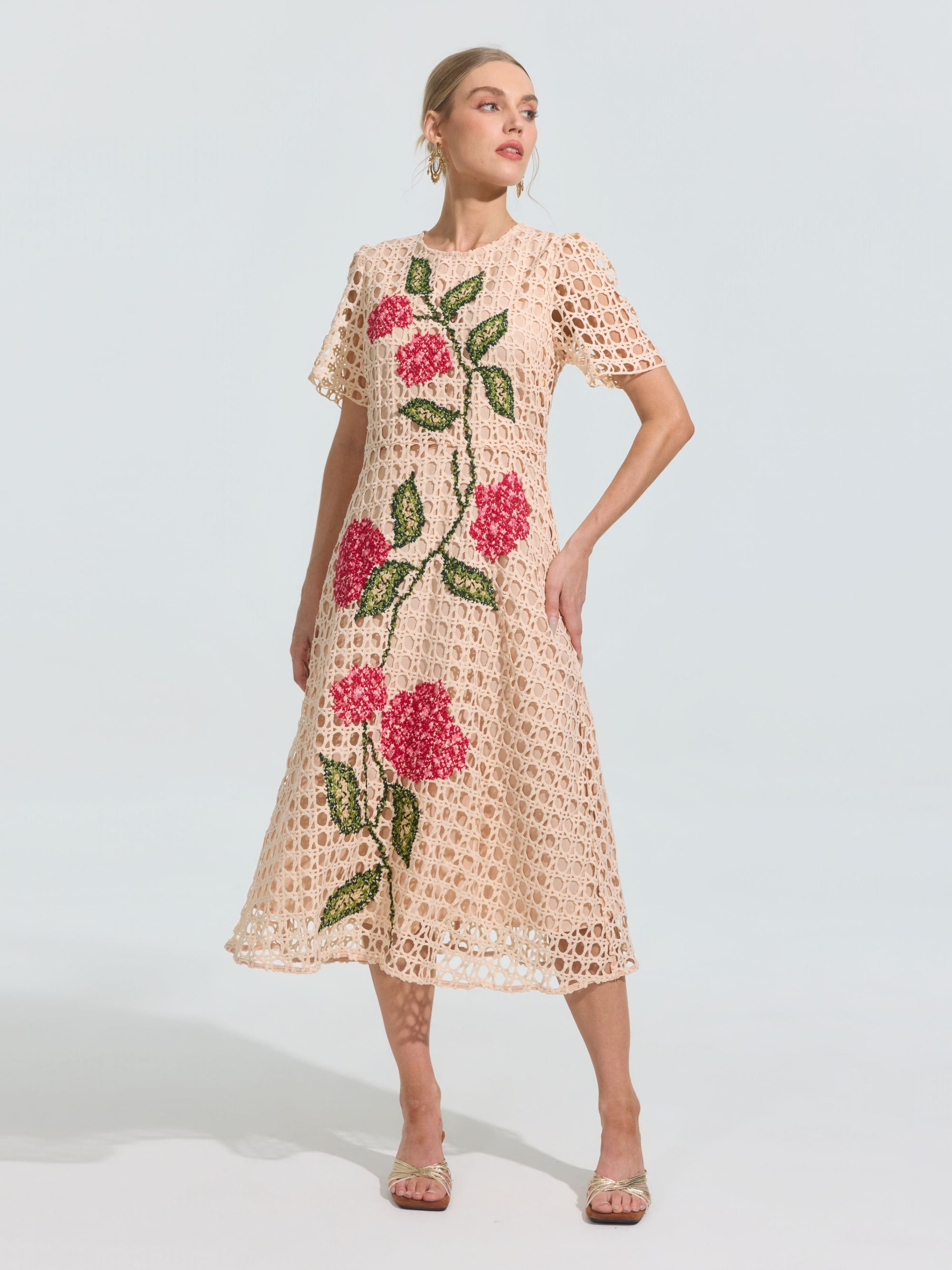 A woman models the Social Queen Hydrangea Embroidered Openwork Dress, a knee-length, short-sleeve beige A-line dress with geometric cutouts and large pink hydrangea and green leaf patterns, set against a plain background.